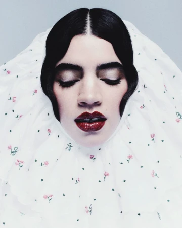 Headshot portrait of a serene woman with closed eyes, dark hair, and red lipstick. She is framed by a white floral-patterned garment.