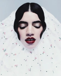 Close-up portrait of a woman with closed eyes wearing a ruffled white floral hood, styled black hair, and glossy dark red lipstick.
