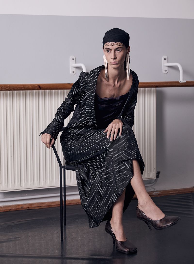 Woman in black dotted suit sits in a dance studio. She wears a black cap and long earrings, with ballet barres in background.