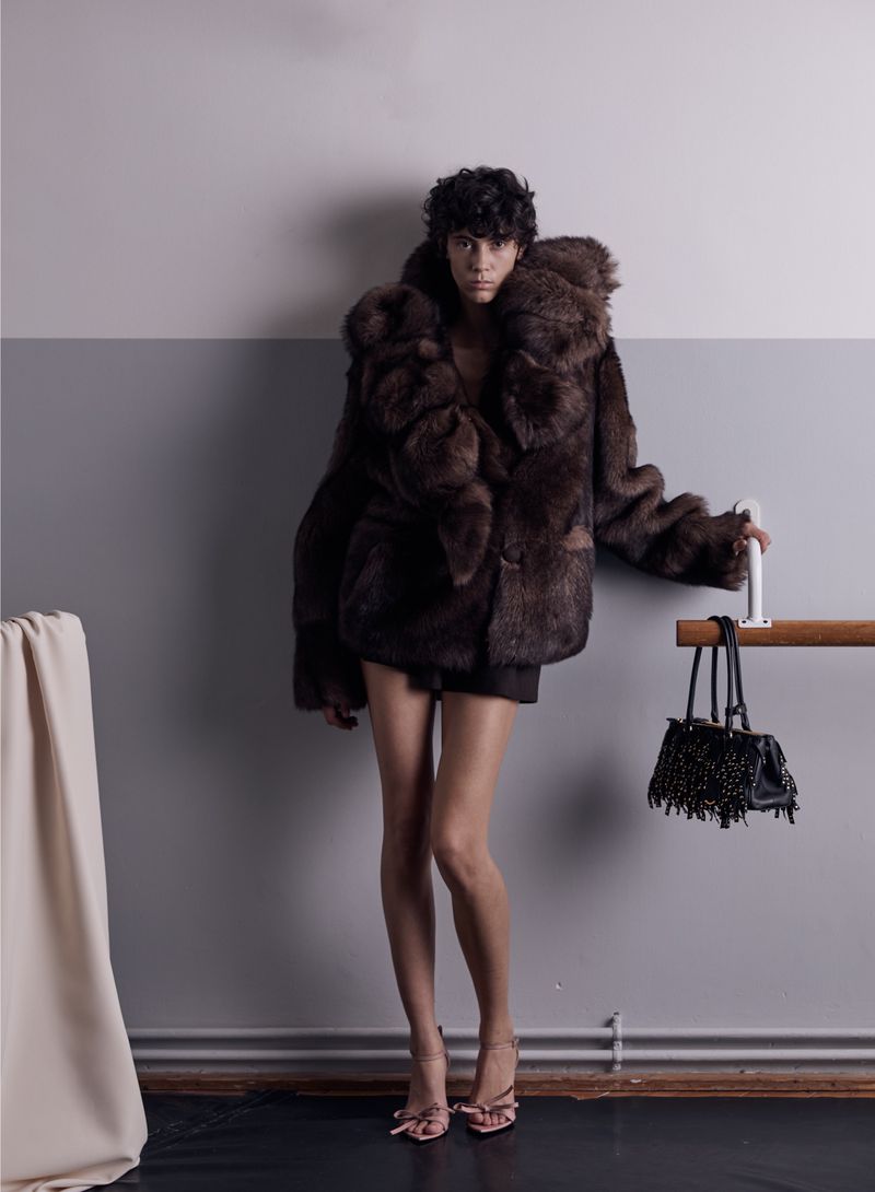 A slender adult female poses in a large brown fur coat, black shorts, and high heels in a minimalist studio with gray and white walls.
