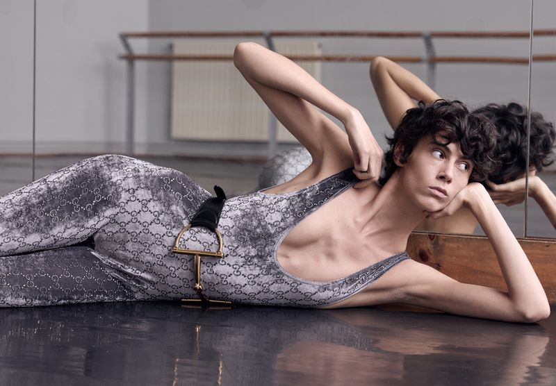 Adult laying on a shiny floor in a patterned bodysuit, with a mirror behind. Fashion and fitness photo shoot in a dance studio.