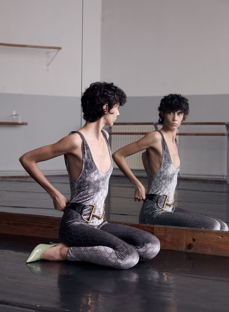 Woman in a snake-print bodysuit and lime-green heels kneeling in a dance studio by a mirror, adjusting her belt and looking into the camera.