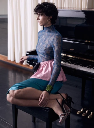 Brunette woman in a semi-sheer lace top with pink and blue skirt sitting on a piano bench with a black piano in the background.