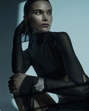Glamour headshot of a woman in black wearing a silver watch, against a cool-toned backdrop with window light. Fashion and beauty portrait.