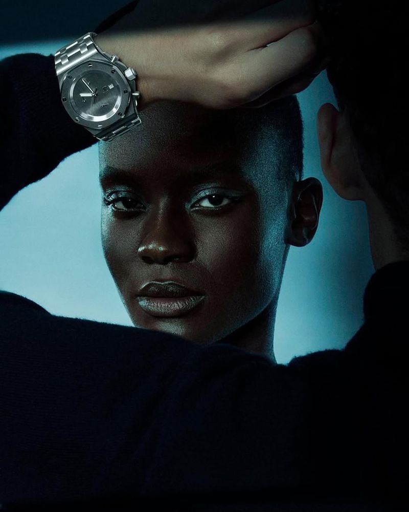 Close-up of an African American woman with short hair and a black top wearing a silver watch on her wrist, set against a light blue background.