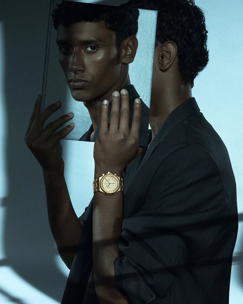 Portrait of a dark-skinned man with a wristwatch, reflected in a mirror, wearing a black shirt against a light blue background.