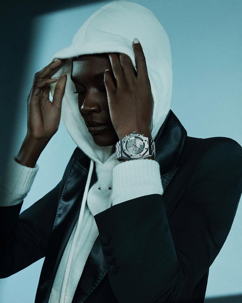A model with dark skin poses indoors wearing a white hooded sweater, black jacket, and a silver watch against a blue background.