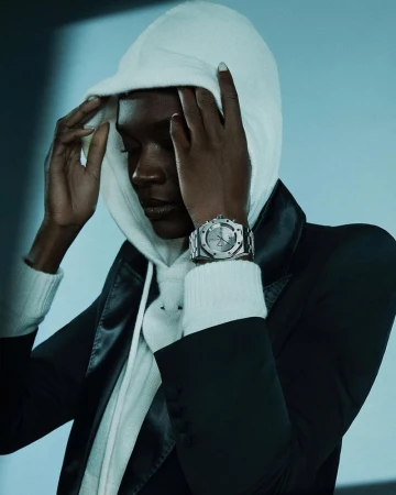 A model with dark skin poses indoors wearing a white hooded sweater, black jacket, and a silver watch against a blue background.