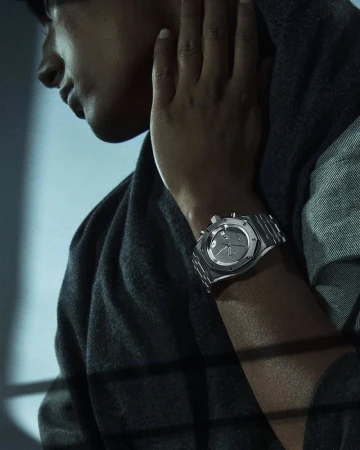 Close-up of a model showcasing a silver wristwatch with a matching bracelet, set against a studio background with soft lighting and shadows.