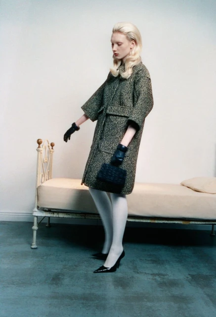 Fashion model poses indoors near vintage bed wearing patterned coat, gloves, tights, heels, and carrying a dark purse in studio lighting.