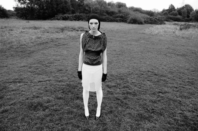 Black and white fashion portrait of a woman in a polka dot top, white skirt, gloves, and beanie standing in a grassy field.