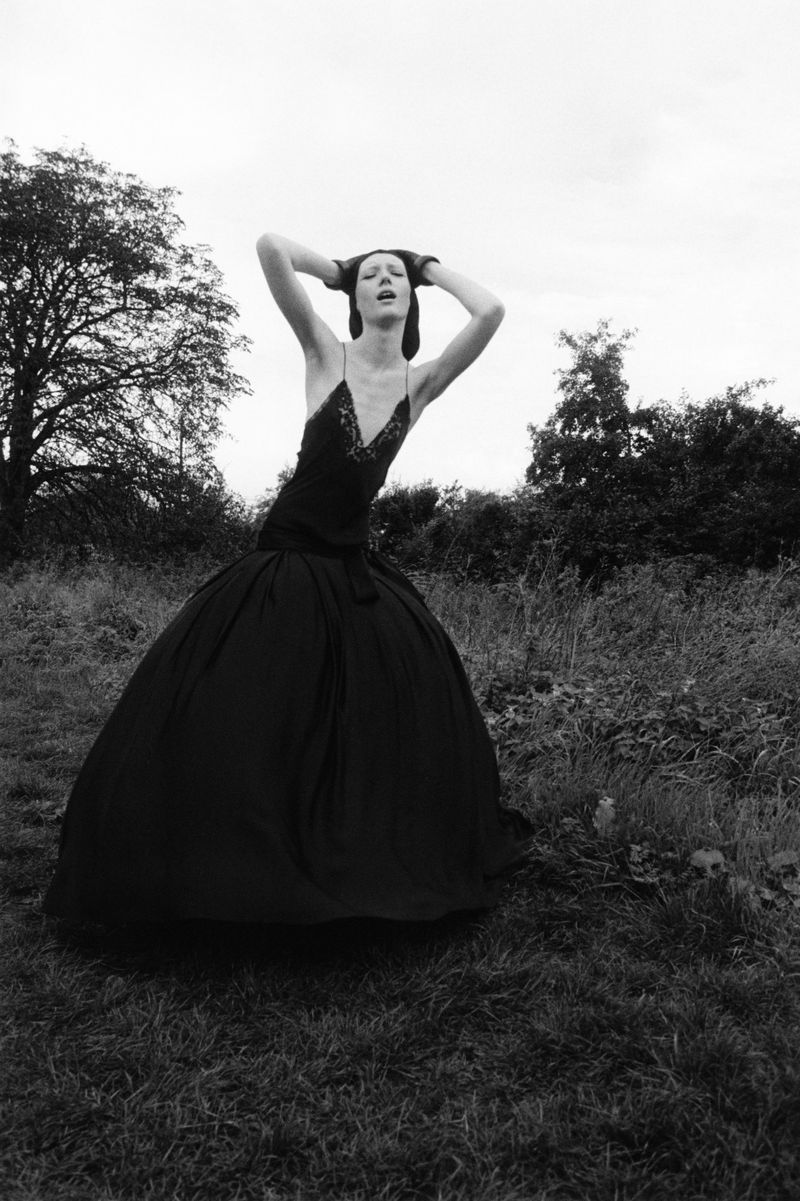 Black-and-white outdoor fashion portrait of a slender female model in a dramatic gown with raised arms, posing in a field.