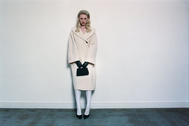 A blonde woman in a beige coat stands in a studio holding a black purse, looking directly at the camera with a neutral expression.