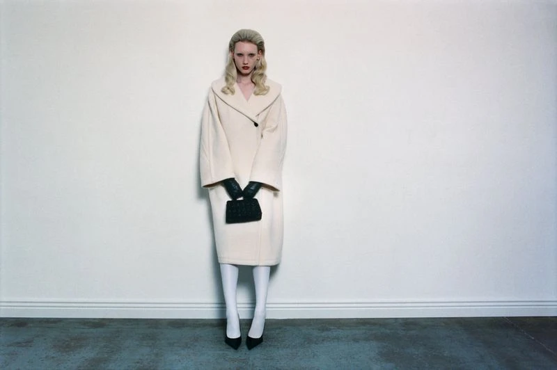 A blonde woman in a beige coat stands in a studio holding a black purse, looking directly at the camera with a neutral expression.