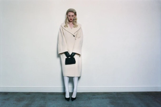 A blonde woman in a beige coat stands in a studio holding a black purse, looking directly at the camera with a neutral expression.