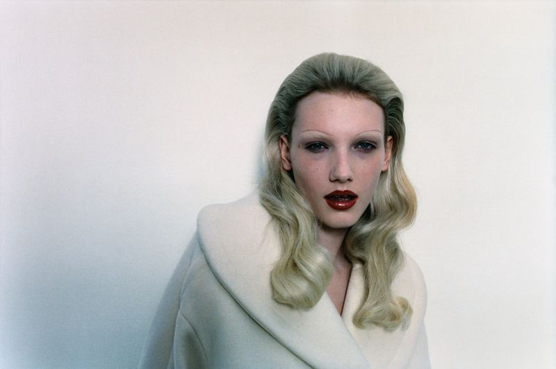 Fashion portrait of a fair-skinned female model with platinum blonde wavy hair, wearing a white coat against a plain white backdrop.