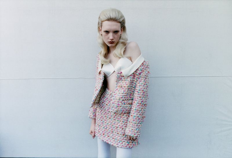 A young adult female model stands in front of a light grey wall wearing a tweed skirt suit with a white top and tights.