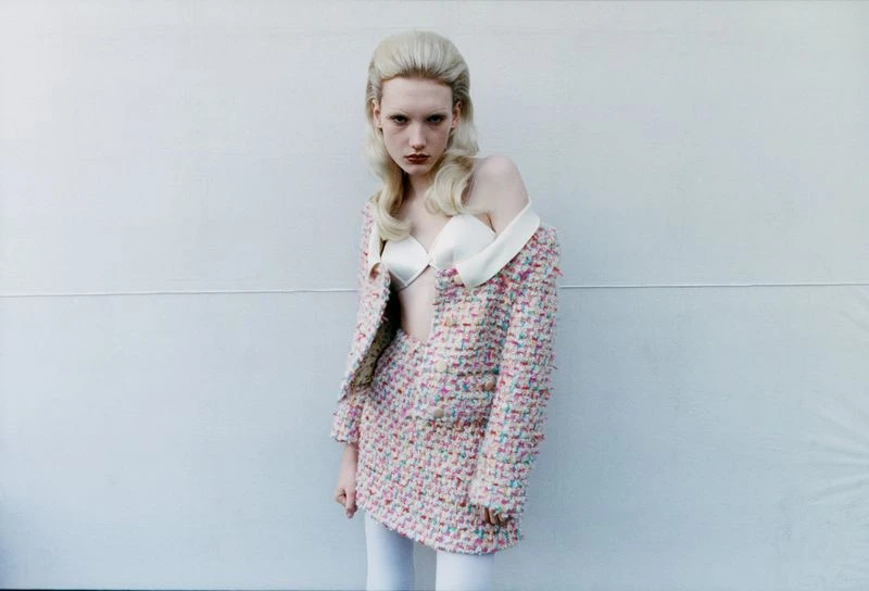 A young adult female model stands in front of a light grey wall wearing a tweed skirt suit with a white top and tights.