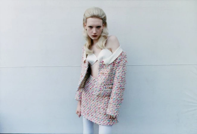 A young adult female model stands in front of a light grey wall wearing a tweed skirt suit with a white top and tights.