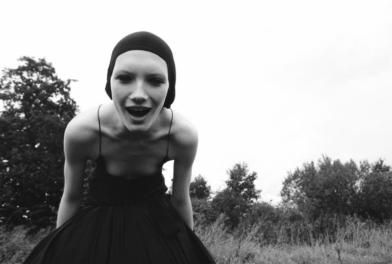 Woman with open mouth wearing a black head covering and dress in a field with trees under a cloudy sky in a black and white photograph.