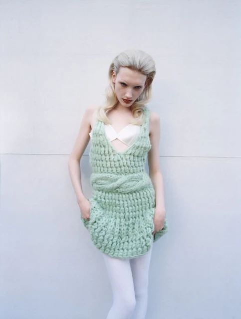 Woman in light green knit dress with hands in pockets posing against white wall in a studio with soft, diffused lighting.