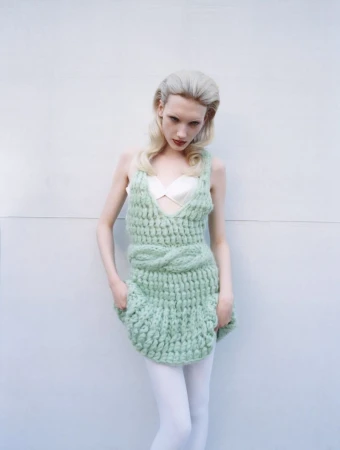 Woman in light green knit dress with hands in pockets posing against white wall in a studio with soft, diffused lighting.