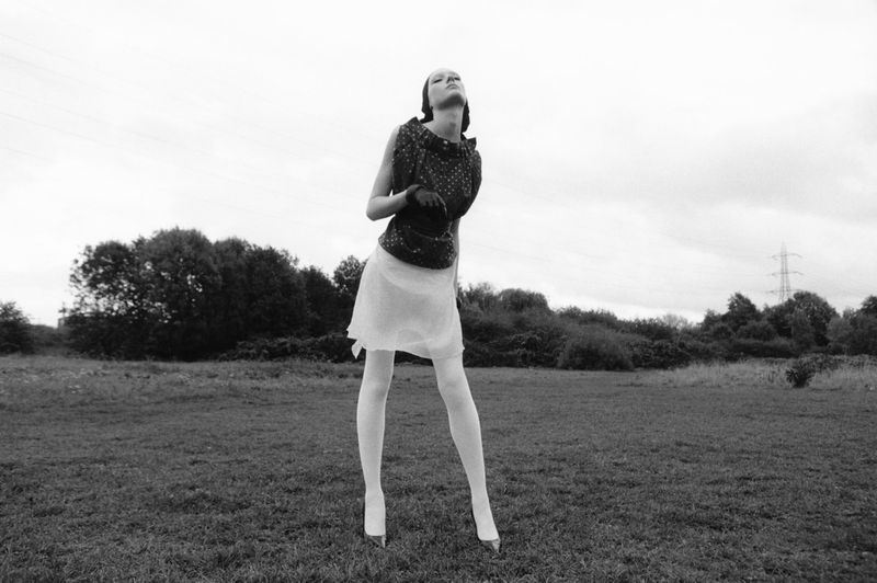 Black and white fashion shot of a woman in a patterned top and skirt, outdoors on a grassy field with trees and cloudy skies.