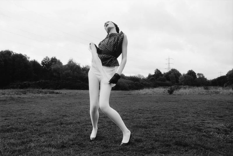 Black and white fashion photograph of a woman in a sleeveless top and white bottoms striking a pose in a field under a cloudy sky.