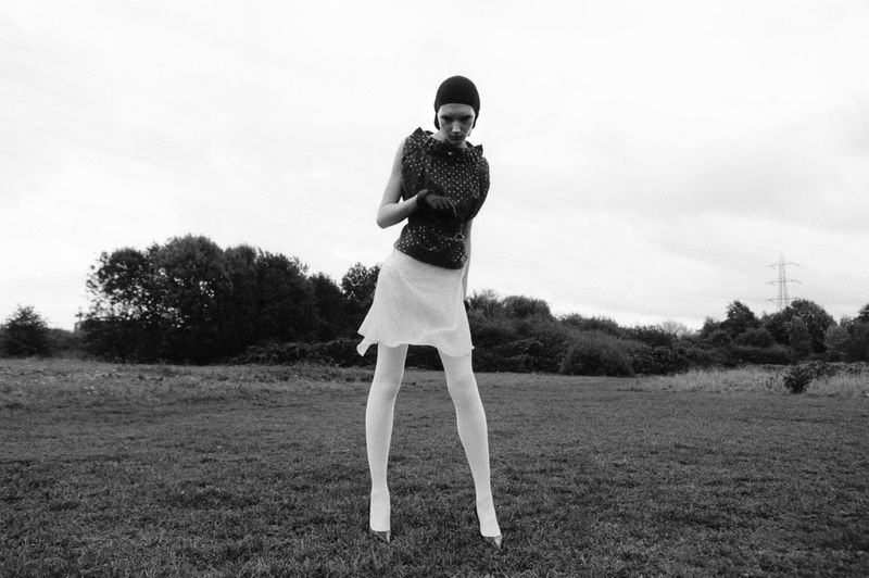 Model Ivy Stewart posing in an open field wearing a dark patterned top, white skirt, white tights, and a dark headpiece.