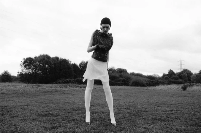 Model Ivy Stewart posing in an open field wearing a dark patterned top, white skirt, white tights, and a dark headpiece.