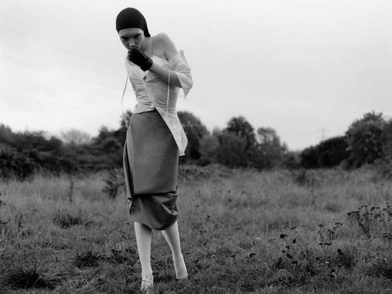 Monochrome image of a person in avant-garde fashion standing in a grassy field, wearing a cap, gloves, and a deconstructed shirt and skirt.