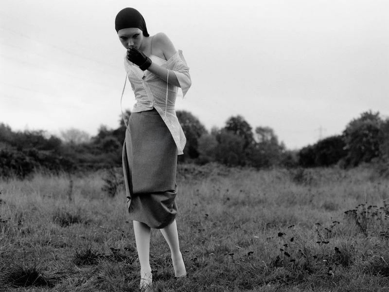 Monochrome image of a person in avant-garde fashion standing in a grassy field, wearing a cap, gloves, and a deconstructed shirt and skirt.