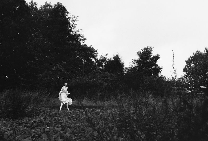 Model Ivy Stewart in a white dress walking through a dark overgrown field framed by tall trees in black and white.