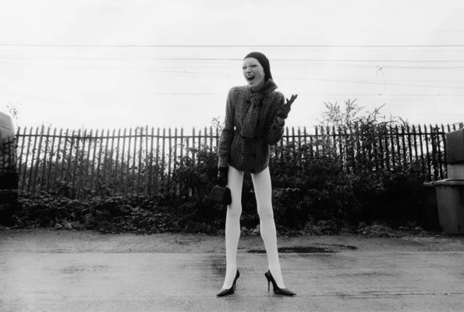 A female fashion model wearing a coat, hat, gloves and heels is standing outdoors in front of a fence in a black and white fashion shot.