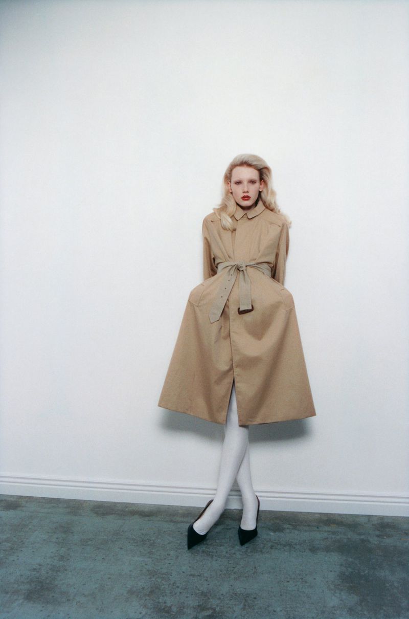 Model in a belted tan coat and white tights standing against a white wall for The Travel Almanac project Between the Marshes.