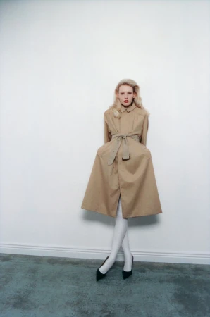 Model in a belted tan coat and white tights standing against a white wall for The Travel Almanac project Between the Marshes.