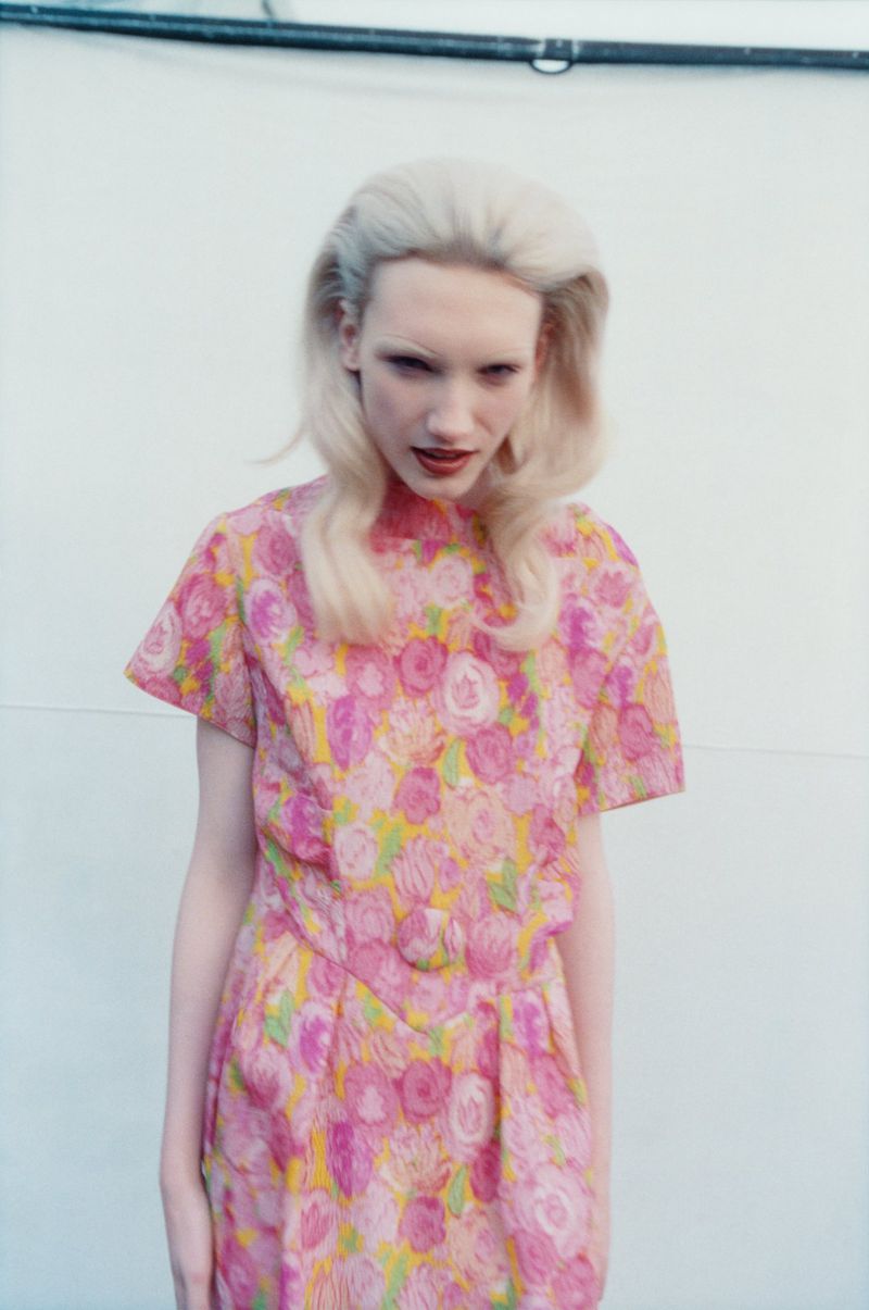 Woman in a floral dress with blonde hair poses against a white wall, portrait-style in medium shot with soft lighting, fashion photography.