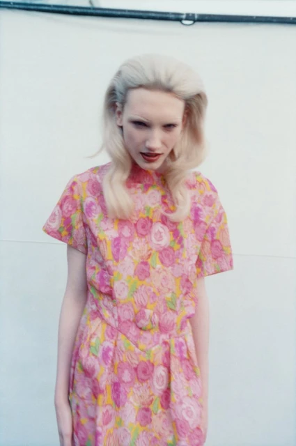 Woman in a floral dress with blonde hair poses against a white wall, portrait-style in medium shot with soft lighting, fashion photography.