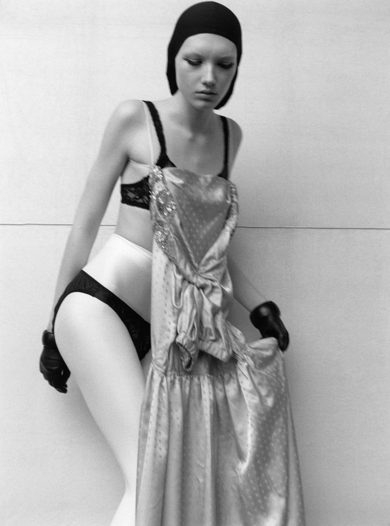 Black-and-white fashion shot of a slim adult woman in lingerie and cap, holding a formal dress in front of a neutral background.