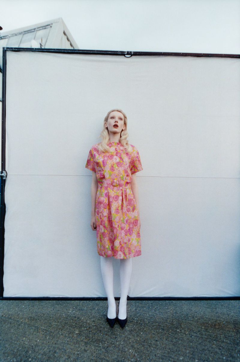 A fair-skinned woman in a pink floral dress stands centered against a white background, wearing black shoes and white tights outdoors.