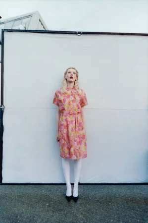 A fair-skinned woman in a pink floral dress stands centered against a white background, wearing black shoes and white tights outdoors.
