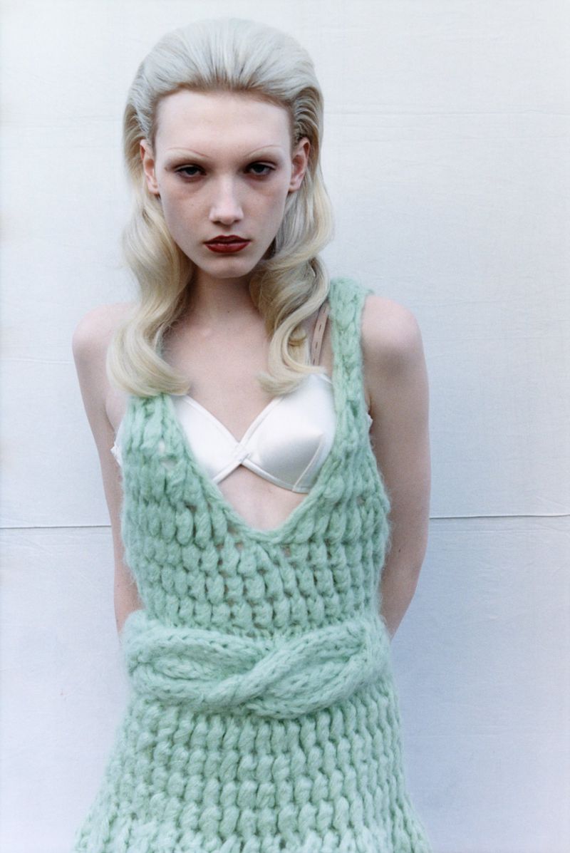 Close-up of a slender young adult female in a light green knitted dress and white lingerie top, posing against a white background. 