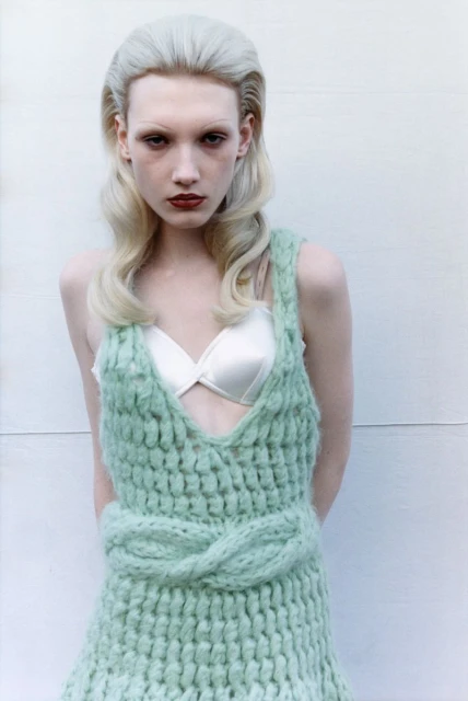 Close-up of a slender young adult female in a light green knitted dress and white lingerie top, posing against a white background. 