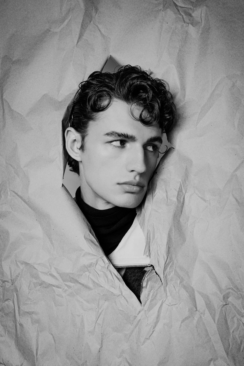 Striking black and white portrait of a dark-haired man with stylish curls, framed by textured white paper in a photo studio setting.