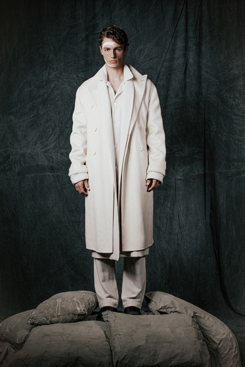Full-length studio shot of a male fashion model wearing an off-white overcoat, shirt, and trousers, standing on a pile of large cushions.