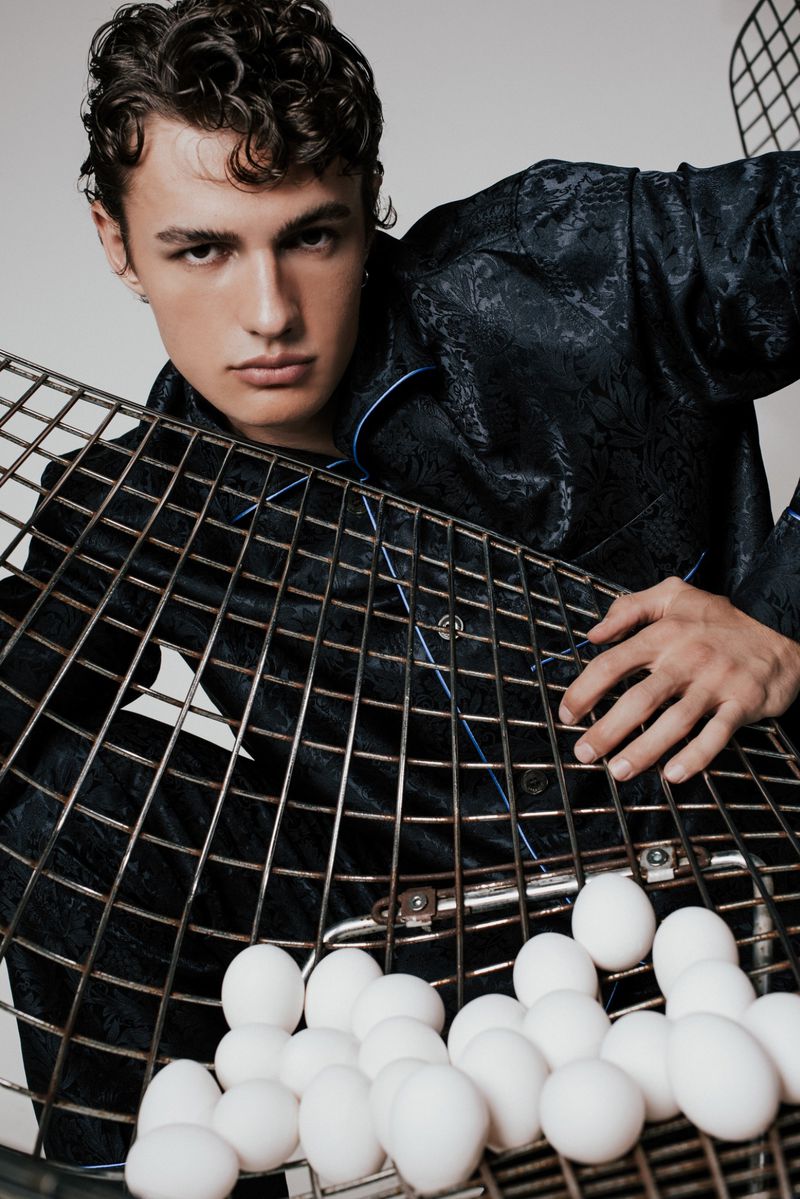 Young man in a patterned pajama top holds a metal wire basket filled with white eggs in a studio shot, looking towards the camera.