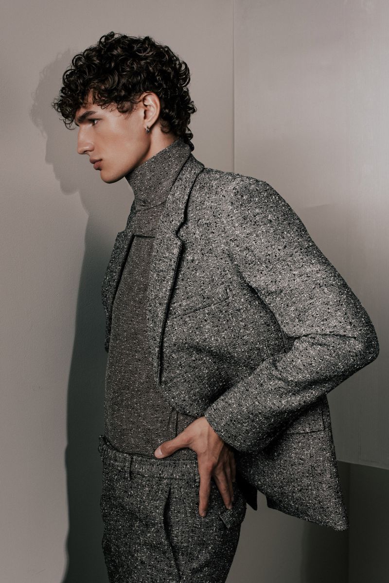 Portrait of a young adult male fashion model with curly hair, wearing a grey patterned suit and turtleneck, standing in a photo studio.