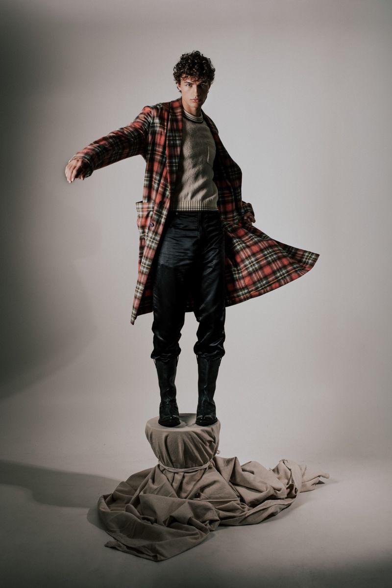 A man wearing a plaid coat and boots stands on a draped pedestal in a studio, posing with open arms against a neutral background.