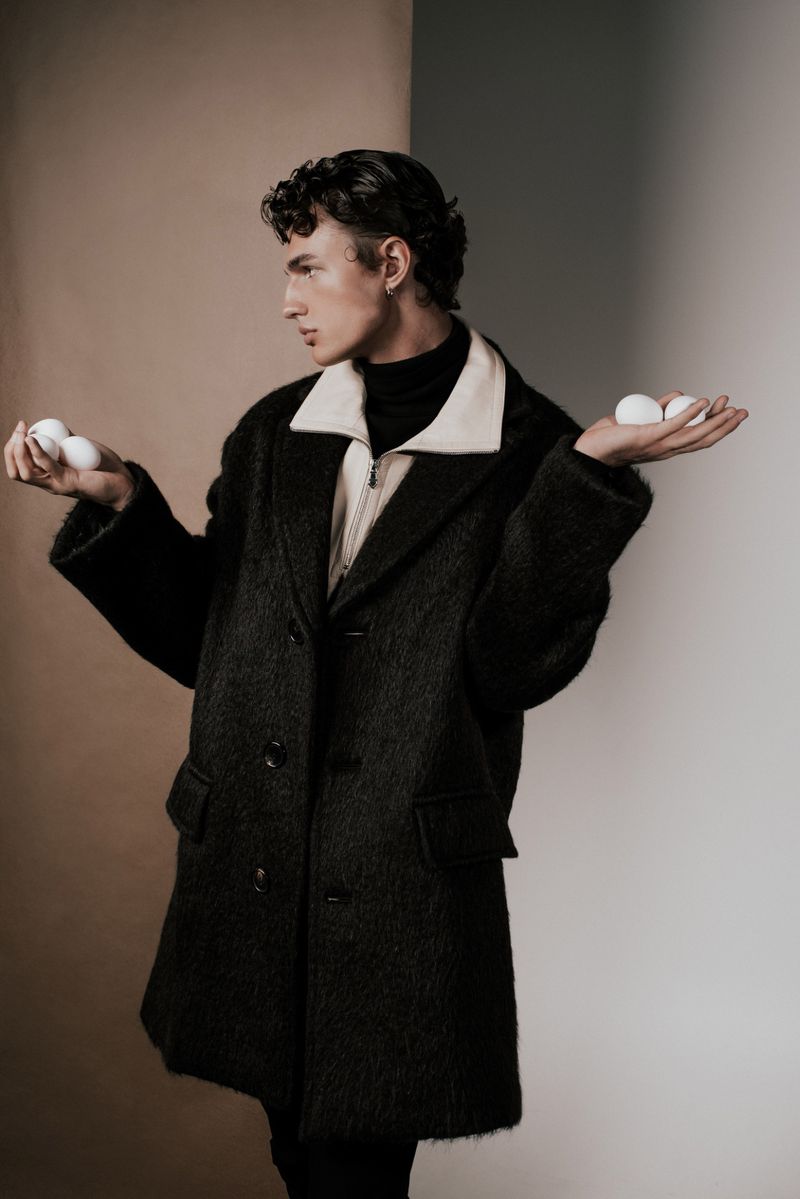 A young adult male is holding eggs in each hand in a studio shot, wearing a dark coat and turtleneck against a split background.
