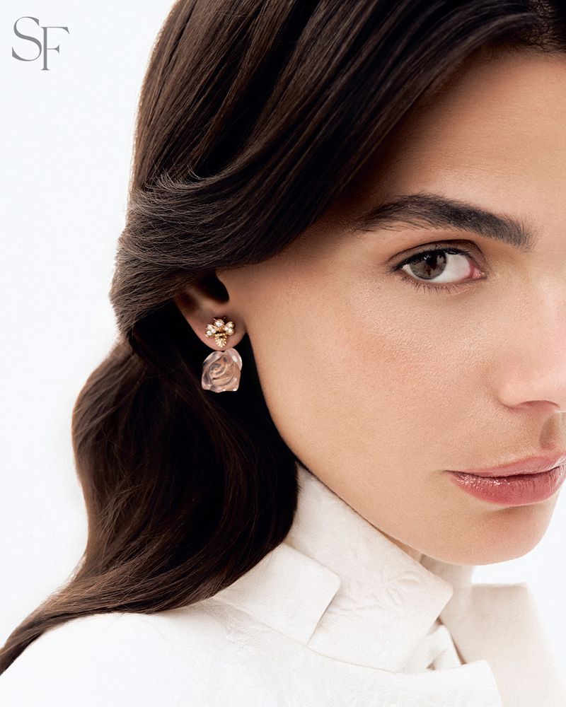Close-up of a woman with dark hair wearing floral-inspired drop earrings and a white jacket against a plain, light-colored background.